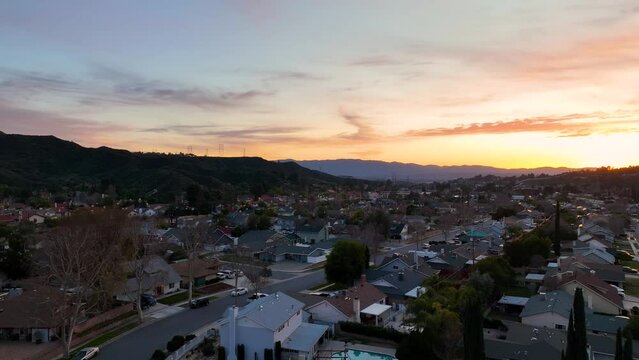 Aerial camera passes over suburb homes in the city of Santa Clarita, Ca. at sunset showing rows of homes and streets with mountains in the background.