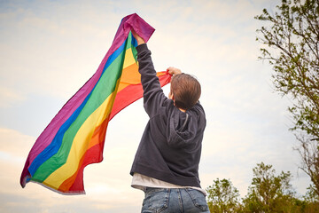 Unrecognizable gay person waving the rainbow flag over the sky, back view. Non-binary person celebrating pride month