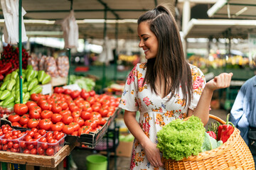 The girl strolls through the market, between stalls with a wooden basket in hands overflowing with an array of fruits and vegetables she has purchased.