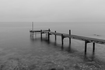 Grayscale shot of a wooden pier on a rocky shore against cloudy sky in Denmark