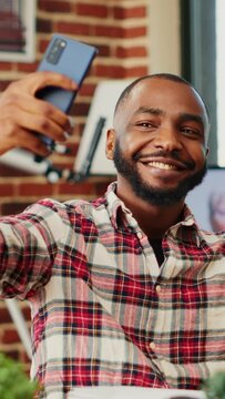 Joyful cheerful african american man taking selfie while smiling and showing thumbs up sign, buttoning up shirt. Person in modern house capturing photo using smartphone with background tv noise
