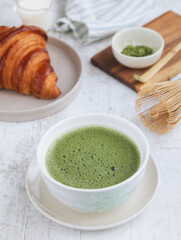 A bowl of traditional Japanese hot matcha green tea with its powder and chesen whisk on a wooden table, with French croissant in the background. A healthy antioxidant food and drink concept 