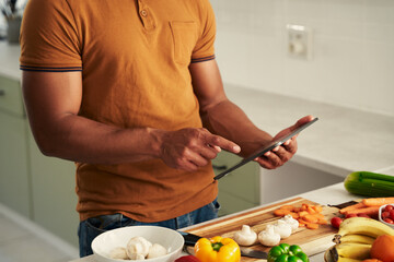 Obraz na płótnie Canvas Close-up of young biracial man in casual clothing using digital tablet next to food in kitchen