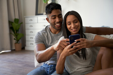 Happy young biracial couple in casual clothing using mobile phone in bedroom at home - Powered by Adobe