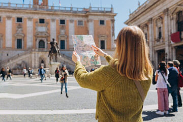 Blonde Woman with paper map is walking on a sunny day. Piazza del Campidoglio on Capitoline Hill, Rome, Italy. Concept of travel, tourism and vacation in city.