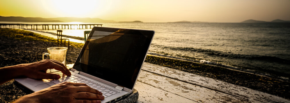 Working on laptop on beach on wooden table at sunset. High resolution photo image can be used as large printed canvas, website banner, social media post. Empty blank copy space for advertising texts.