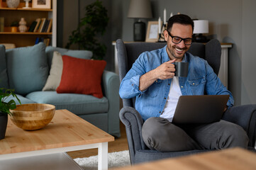 Smiling young businessman drinking coffee and reading e-mails over wireless computer while sitting...