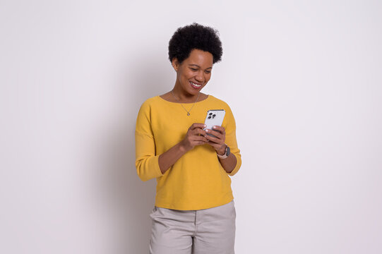 Smiling Young Woman With Short Afro Hair Text Messaging On Smart Phone. Beautiful Female Dressing In Casuals Using Social Media Over Cellphone While Standing Isolated On White Background