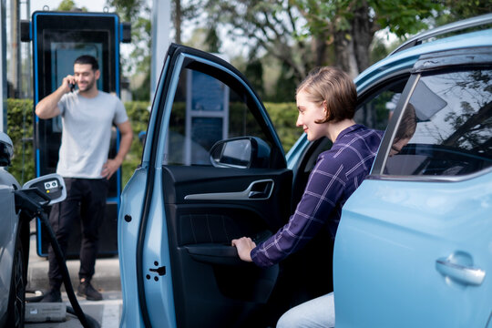 Rusain woman opend car's door while her boyfriend talking on his smartphone with smile while waiting for fill green energy by save money with electral car fluel station with blur happy her boyfriend.