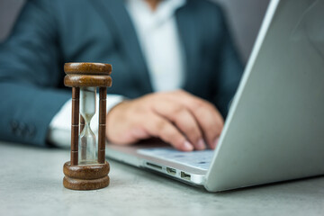 Businessman typing on a laptop keyboard next to a wood hourglass. Concept of time management, business schedule, and deadline, time concept
