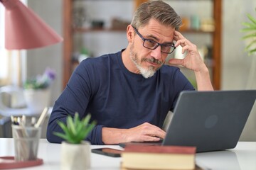Casual mid adult man with laptop computer at desk in home office, banking online, remote working. Portrait of older gray haired bearded guy thinking. Businessman managing business on internet.  