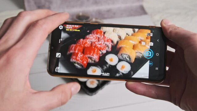 A male hands take a photo of sushi rolls in a delivery plastic box with a smartphone camera. Blogger shoots a review of Japanese food on a mobile phone for posting on social networks. Selective focus