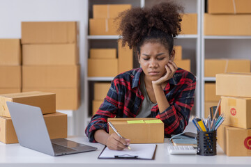 
Portrait of Young African American business woman SME e-commerce employee sitting at home full of packages write note of orders use a calculator and laptop online SME business idea concept