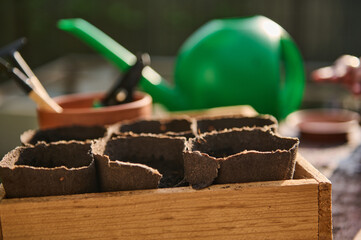 Still life with disposable peat pots on wooden crate against blurred green watering can and garden...