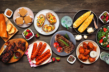 Summer BBQ food table scene over a dark wood background. Assorted grilled meats, potatoes,...