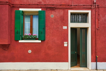 Fototapeta na wymiar Door and window with flower on the red facade of the house. Colorful architecture in Burano island, Venice.