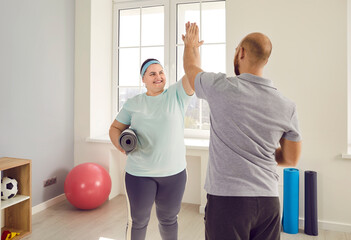 Happy, fat, large, overweight, young woman with exercise mat and man who works as her personal sports trainer give each other high five after successful fitness workout at gym - Powered by Adobe