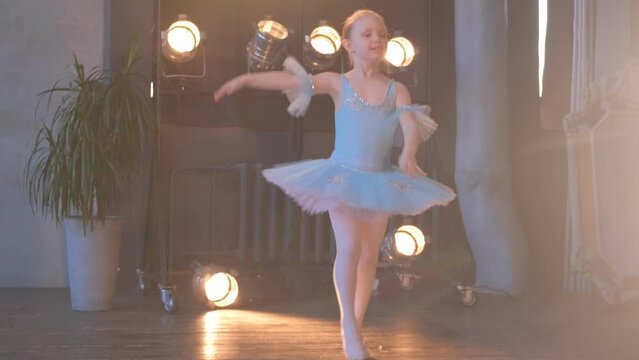 Adorable Girl Ballerina In White Suit With Hair Bun On Head Does Exercise For Hand In Brightly Lit Studio At Dance Lesson.