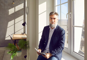 Bearded middle aged gray haired man sitting by window in office. Portrait of handsome confident...