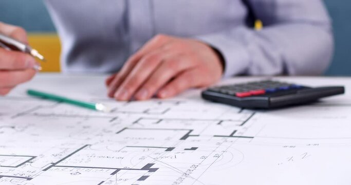 Engineer in office clothing makes calculations using calculator. Architect leaves notes with lead pencil on building drawing while sits at table