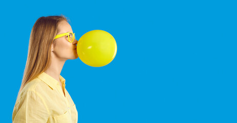 Happy student girl inflating balloon for fun party games. Profile portrait of long haired young woman in glasses blowing up yellow balloon standing on empty blank blue text copyspace banner background