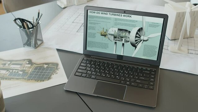 Close-up shot of desk of architectural engineer at energy efficient home construction company, containing laptop with wind turbine design on screen, blueprints, plans and small scale house models