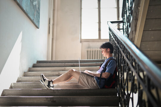 young man sitting on the stairs of a school uses a computer
