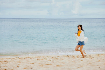 Sports woman runs along the beach in summer clothes on the sand in a yellow T-shirt and denim shorts white shirt flying hair ocean view