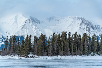 Winter season views in northern Canada with snow capped towering mountain landscape, boreal forest, spruce, pine trees, frozen lake in foreground. 