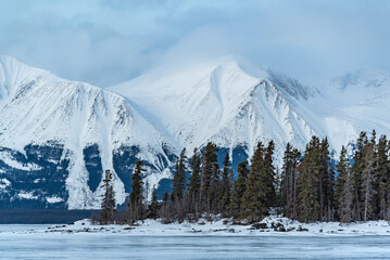 Winter season views in northern Canada with snow capped towering mountain landscape, boreal forest, spruce, pine trees, frozen lake in foreground. 