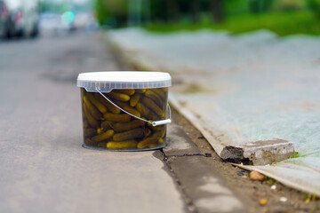 A plastic transparent bucket of pickles stands on the sidewalk