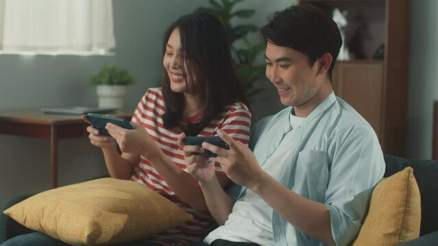 Portrait of happy young attractive Asian couple hand holing mobile phone enjoy playing MMORPG or MOBA game on smartphone feeling cheerful fun and excited sit on couch at cozy home living room
