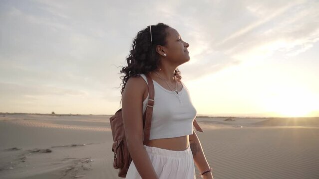 Smiling African American woman standing peacefully on desert dunes summer day. Curly pretty girl relaxing on sand. Happy young black people enjoying leisure in nature. Vacations slow motion. 