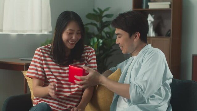Happy attractive Asian couple giving gift box surprise to celebrate anniversary day or birthday. Attractive Young Man hug his girlfriend with love sit on couch at living room. Lover lifestyle concept