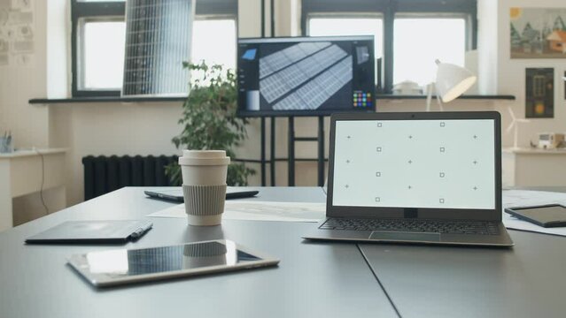 Full shot of laptop with white screen on desk of building engineer at green energy construction company, plans, solar panels, small scale models of houses and wind turbine. Template, copy space