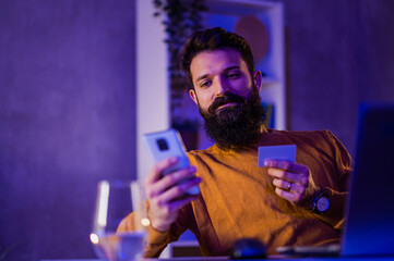 A hipster is using his credit card and smartphone for online shopping while sitting at his neon blue and purple lighted home office.