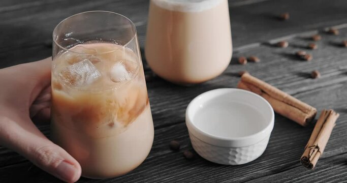 Iced latte drinks for two, person placing a glass with coffee cocktail with milk froth and ice cube on wooden table decorated with coffee beans and cinnamon sticks, 4k close up footage, horizontal