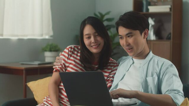 Portrait of happy young attractive Asian couple using laptop computer enjoy playing social media surfing internet watching live steam or online movie smiling sit on couch at cozy home living room