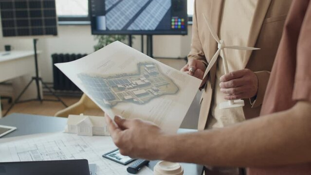 Close-up shot of hands of unrecognizable male architects standing around desk in renewable energy startup office, holding wind turbine and plan of energy efficient buildings complex and arguing