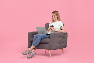 Fototapeta na wymiar Happy woman with laptop and cup of drink sitting in armchair against pink background