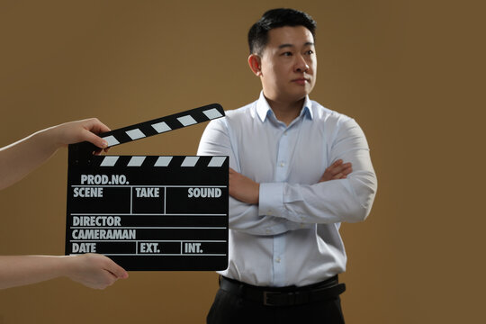Asian actor performing while second assistant camera holding clapperboard on brown background, selective focus. Film industry