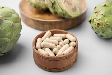 Bowl with pills and fresh artichokes on light grey background, closeup