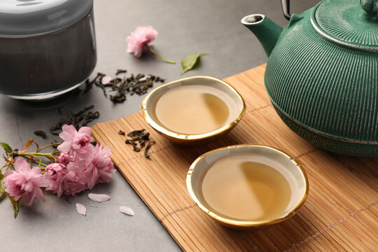 Traditional ceremony. Cups of brewed tea, teapot and sakura flowers on grey table, closeup