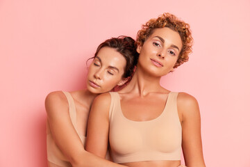 Two young women in beige tops stand on pale pink studio background, one hugging the curly...