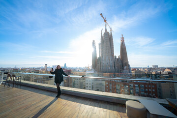 Young woman traveler in outerwear standing on balcony and taking selfie on smartphone against famous building Sagrada Familia basilica by Gaudi, during sightseeing trip in Barcelona Spain in sunlight