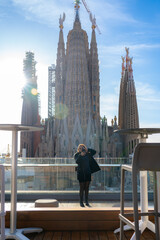 Young woman traveler in outerwear standing on balcony and taking selfie on smartphone against famous building Sagrada Familia basilica by Gaudi, during sightseeing trip in Barcelona Spain in sunlight