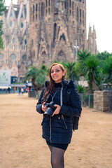 Serious young Latin woman tourist in warm outfit with photo camera looking at camera while standing against amazing church Sagrada Familia by Gaudi during sightseeing trip through Barcelona, Spain