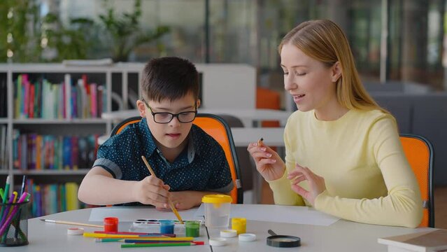 Adorable boy with Down syndrome having art class with his teacher, drawing picture with watercolors