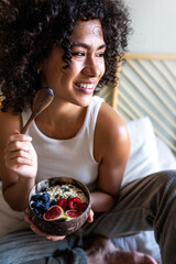 Vertical portrait of happy young latina woman smiling eating healthy breakfast bowl of oats and fruit sitting on bed.