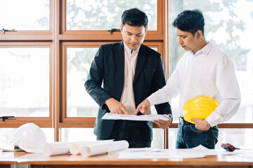 Two Asian male architectural teams, wearing uniforms, point to blueprints working together on engineering projects. Construction industry business plan in office with safety helmet